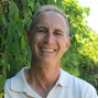 Head-and-shoulders portrait of Scott Thomas wearing a white collared shirt, photographed outdoors with green foliage and a wooden structure in the background. 