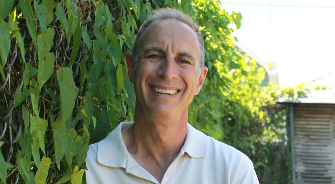 Head-and-shoulders portrait of Scott Thomas wearing a white collared shirt, photographed outdoors with green foliage and a wooden structure in the background.