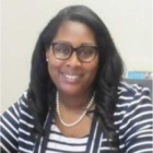 Person with long wavy dark hair wearing a striped top and a pearl necklace, seated indoors against a light‑colored background. 