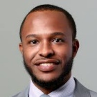 Person wearing a light gray suit with a light blue striped dress shirt and matching tie, posed against a plain gray background. 