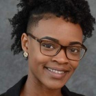 Person with short, curly dark hair wearing small decorative earrings and a dark-colored top, posed against a textured gray background. 