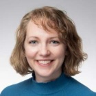 Person with shoulder‑length wavy blond hair wearing a bright blue sweater against a plain light gray background. 