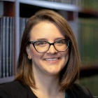 Person with shoulder‑length brown hair wearing a dark blazer, seated in front of shelves filled with books. 