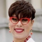Person with short dark hair wearing a white top and red beaded jewelry, standing outdoors in front of a light‑colored building. 