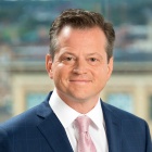 Person wearing a navy blue checkered suit with a white shirt, light pink tie, and white pocket square, standing indoors with an out‑of‑focus cityscape in the background. 