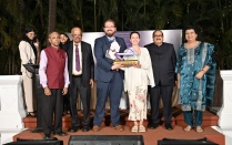 A group of people stand together on an outdoor stage during an awards event. Two people at the center hold a trophy and certificate, with others standing on either side in formal and semi formal attire. 