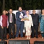 A group of people stand together on an outdoor stage during an awards event. Two people at the center hold a trophy and certificate, with others standing on either side in formal and semi formal attire. 