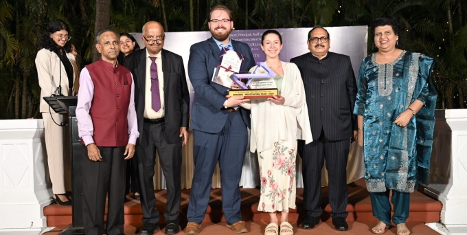 A group of people stand together on an outdoor stage during an awards event. Two people at the center hold a trophy and certificate, with others standing on either side in formal and semi‑formal attire. 