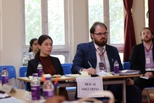 Three participants sit at a table labeled ‘Med AI Solutions’ during an indoor event, taking notes and listening. Water bottles, papers, and pens are on the table, and large windows with curtains are in the background. 