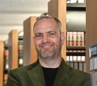 A person standing in a library aisle with bookshelves behind them, wearing a dark jacket. 