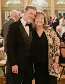 A couple dressed in formal evening attire stands together in an elegant ballroom with arched windows and ornate columns. Other guests sit and mingle at tables in the background during what appears to be a formal event or celebration. 