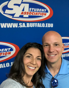 A couple poses in front of a blue backdrop featuring the UB Student Association logo and the website 'www.sa.buffalo.edu.' One person wears a light blue collared shirt with sunglasses hanging from it, and the other has long dark hair. The backdrop repeats the Student Association branding. 