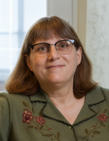 A person with shoulder‑length light brown hair sits indoors near a bright window. They are wearing an olive‑green cardigan decorated with embroidered red and pink flowers and leafy vines along the neckline and chest. 
