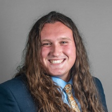 Professional portrait of a person in a navy suit, light blue shirt, and floral patterned tie against a gray background. 