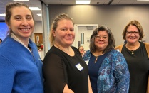 Four adults stand together indoors, smiling and conversing, wearing business casual attire in a hallway or event space. 