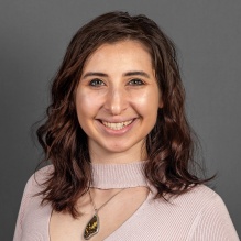Studio portrait of an adult wearing a light colored top and a pendant necklace, standing against a plain gray background. 