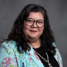 Studio portrait of an adult wearing a black top, a floral patterned jacket, and a long necklace, posed against a gray background. 