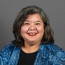 Studio portrait of an adult wearing a black top, a floral patterned jacket, and a long necklace, posed against a gray background. 