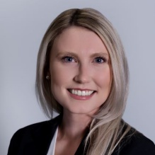 Studio portrait of an adult wearing a dark blazer over a light blouse, with long light colored hair, photographed against a plain light background. 