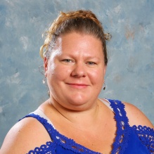 Studio portrait of an adult wearing a blue sleeveless top with lace detail, photographed against a mottled blue background. 