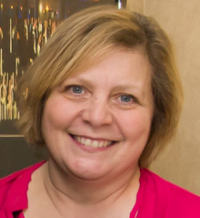 Indoor portrait of an adult wearing a bright pink top, with short light colored hair, standing in front of a framed artwork on a wall. 