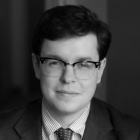 Black-and-white professional portrait of a person wearing glasses, a suit jacket, striped shirt, and patterned tie, seated indoors beside a table lamp. 