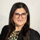 Studio portrait of a person with dark hair and glasses, wearing a black blazer over a floral patterned top against a light background. 