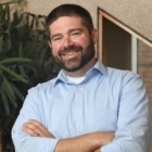 Close‑cropped professional portrait of a person wearing a light blue shirt, photographed indoors against a softly blurred, light‑colored background. 