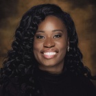 Professional headshot of a person with long curly hair wearing a dark top against a dark brown studio background. 