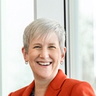 Professional portrait of a person in an orange blazer standing beside floor to ceiling windows in a modern campus building. 