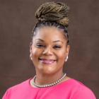 Professional headshot of a person wearing a pink top and pearl jewelry against a brown studio background. 