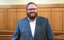 Person in a suit standing inside a courtroom, leaning against a wooden railing with the judge&rsquo;s bench and seating visible in the background. 