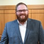 Person in a suit standing inside a courtroom, leaning against a wooden railing with the judge&rsquo;s bench and seating visible in the background. 
