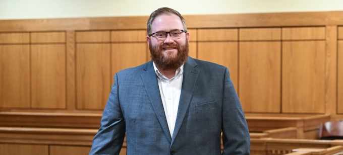 Person in a suit standing inside a courtroom, leaning against a wooden railing with the judge’s bench and seating visible in the background. 