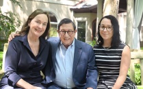 Three people sit side by side on a bench in a garden courtyard with greenery and a building in the background. 