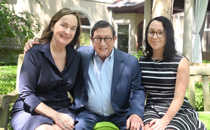 Three people seated together on a wooden bench in an outdoor courtyard surrounded by trees and landscaping. 