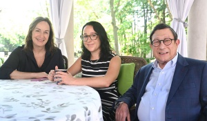Three people sit together at a patio table in an outdoor gathering space with greenery, stone architecture, and large draped curtains in the background. 