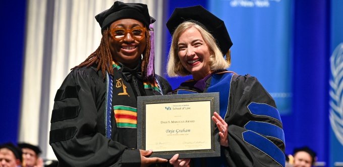Graduation ceremony scene showing two people in academic regalia on stage holding a framed certificate that reads “University at Buffalo School of Law Dale S. Margulis Award Deja Graham,” with an audience seated in the background. 
