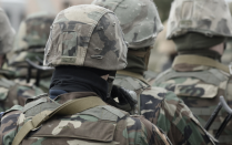 Rear view of several people wearing camouflage military uniforms and helmets standing together outdoors, with the foreground figure centered and the background softly blurred. 
