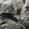 Rear view of several people wearing camouflage military uniforms and helmets standing together outdoors, with the foreground figure centered and the background softly blurred. 