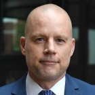 Professional portrait of a person in a blue suit and patterned tie standing outdoors in front of a glass building, with hands clasped and the background softly blurred. 