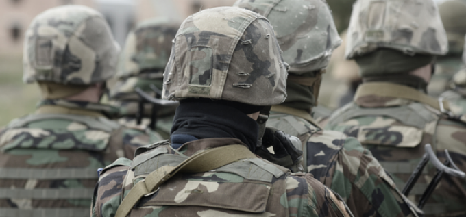 Rear view of several people wearing camouflage military uniforms and helmets standing together outdoors, with the foreground figure centered and the background softly blurred. 