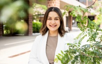 Farina Barth standing outdoors on a campus walkway, wearing a light colored jacket and holding a blue folder, with brick buildings and green foliage visible in the background. 