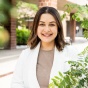 Farina Barth standing outdoors on a campus walkway, wearing a light colored jacket and holding a blue folder, with brick buildings and green foliage visible in the background. 