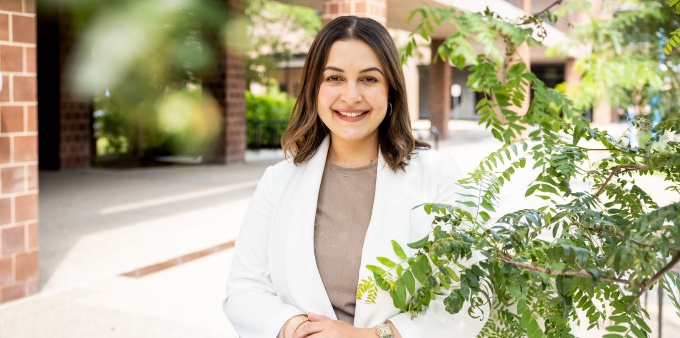 Farina Barth standing outdoors on a campus walkway, wearing a light colored jacket and holding a blue folder, with brick buildings and green foliage visible in the background. 