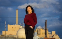 Person standing outdoors in front of an industrial facility with smokestacks, under a cloudy blue sky. 
