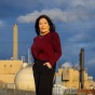 Person standing outdoors in front of an industrial facility with smokestacks, under a cloudy blue sky. 