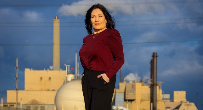Person standing outdoors in front of an industrial facility with smokestacks, under a cloudy blue sky. 