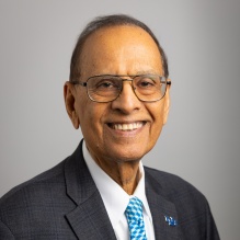 Portrait of Satish Tripathi wearing blue tie, dark grey jacket, white shirt, glasses, in front of a grey backdrop. 