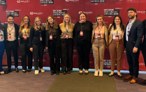 Group of eleven attendees standing shoulder to shoulder in front of a red conference backdrop with event branding, wearing business and business casual attire and conference name badges at an indoor venue. 